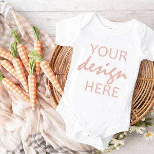 May include: White baby onesie with the text "YOUR design HERE" in pink on a white wicker basket with orange and white checked carrots on a tan and white checked fabric.