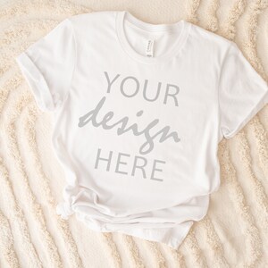 May include: White t-shirt with the text "YOUR design HERE" printed on the front. The shirt is lying on a white fluffy surface.