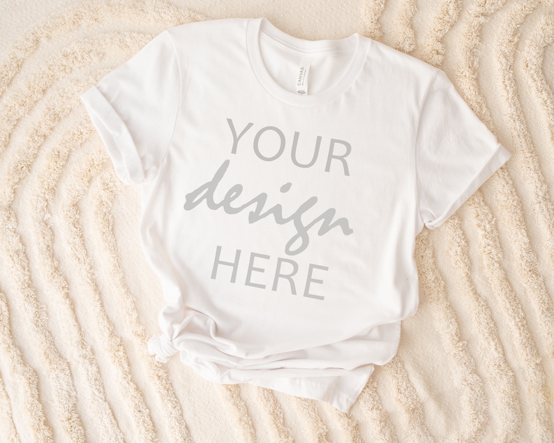 May include: White t-shirt with the text "YOUR design HERE" printed on the front. The shirt is lying on a white fluffy surface.