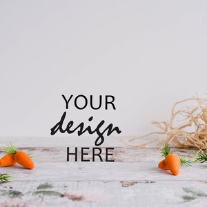 May include: A whitewashed wooden surface with the text "YOUR design HERE" in black.  There are five orange carrots with green stems scattered on the surface.  A pile of straw is in the background.
