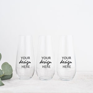 以下が含まれることがあります： 黒い筆記体で「YOUR design HERE」と書かれた、3つの透明なステムレスシャンパングラス。グラスは白い表面に置かれ、白い背景と緑の葉の小枝があります。