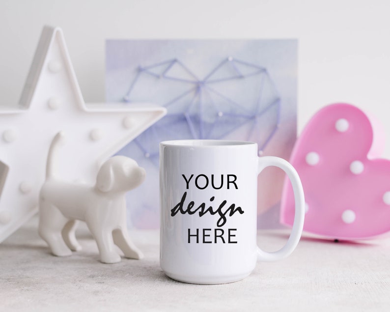 May include: White ceramic mug with a black inscription that reads "YOUR design HERE". The mug is sitting on a white surface with a white star, a white dog figurine, and a pink heart-shaped light in the background.