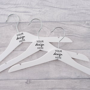 Peut inclure: Trois cintres en bois blanc avec des crochets argentés. Chaque cintre porte l'inscription "YOUR design HERE" imprimée sur le devant.