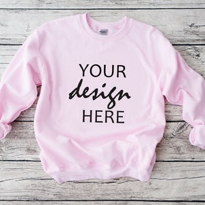 以下が含まれることがあります： 黒字で「YOUR design HERE」と書かれた、明るいピンク色のスウェットシャツ。明るい木目調の表面に表示されています。袖口は折り返されています。