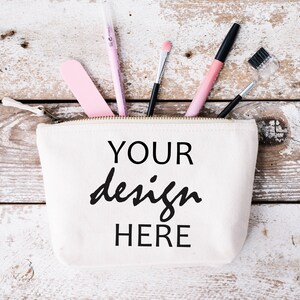 May include: A white canvas makeup bag with a zipper closure. The bag has the text "YOUR design HERE" printed on the front in black.