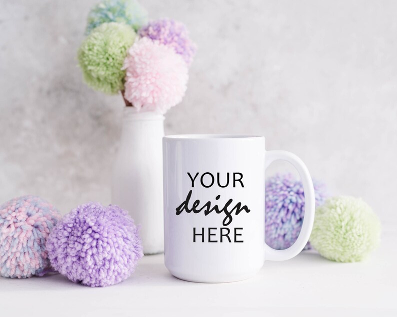 May include: White ceramic mug with the text "YOUR design HERE" in black lettering. The mug is surrounded by colorful pom-poms.