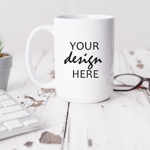 May include: White ceramic mug with a black inscription that reads "YOUR design HERE" on a white wooden surface with a small potted plant and a pair of eyeglasses.