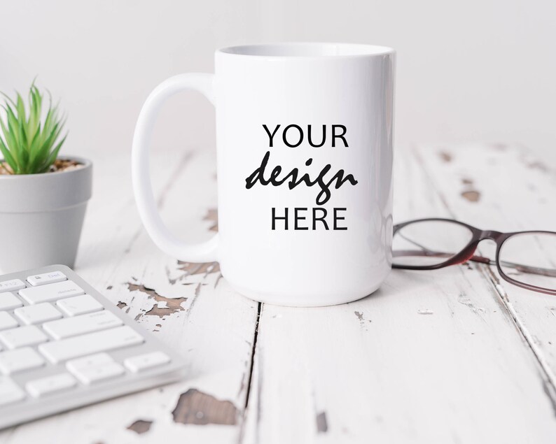 May include: White ceramic mug with a black inscription that reads "YOUR design HERE" on a white wooden surface with a small potted plant and a pair of eyeglasses.