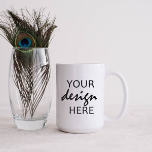 May include: White ceramic mug with a black inscription that reads "YOUR design HERE". The mug is sitting on a white surface next to a glass vase with peacock feathers.