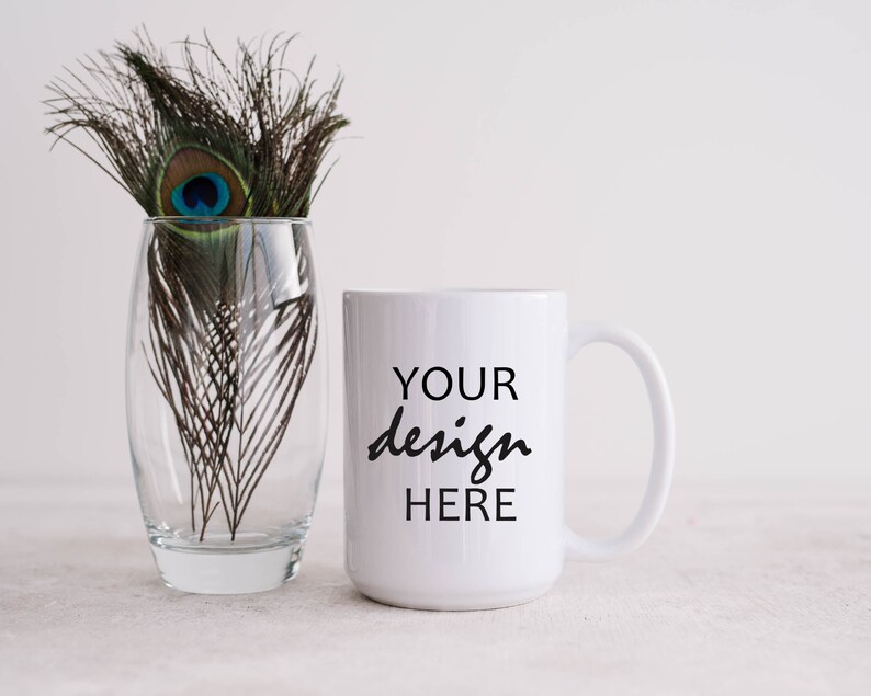 May include: White ceramic mug with a black inscription that reads "YOUR design HERE". The mug is sitting on a white surface next to a glass vase with peacock feathers.