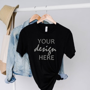 May include: Black t-shirt with the text "YOUR design HERE" hanging on a white rack with a denim jacket and a tote bag.