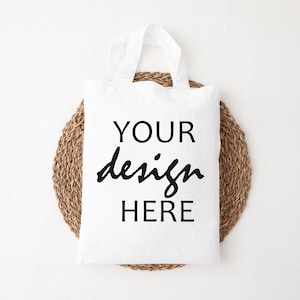 May include: White tote bag with the text "YOUR design HERE" printed on it. The bag is sitting on a round woven mat.