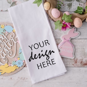Op de afbeelding: Een witte keukendoek met de tekst "YOUR design HERE" in zwart gedrukt. De doek ligt op een wit houten oppervlak met paasversieringen, waaronder een krans met roze en bruine eieren, een houten bord met de tekst "Happy Easter" en een roze konijnenbeeldje met de tekst "Happy Easter" erop.