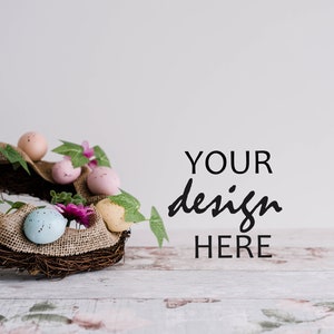 May include: A brown twig wreath decorated with pastel pink, blue, and yellow speckled Easter eggs, green leaves, and pink flowers. The wreath is sitting on a whitewashed wooden surface with the text "YOUR design HERE" in black.