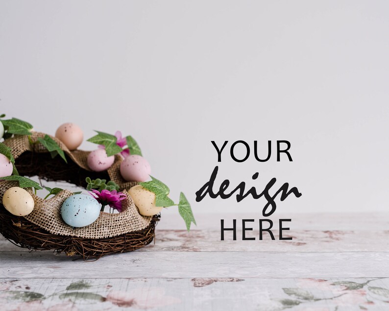 May include: A brown twig wreath decorated with pastel pink, blue, and yellow speckled Easter eggs, green leaves, and pink flowers. The wreath is sitting on a whitewashed wooden surface with the text "YOUR design HERE" in black.