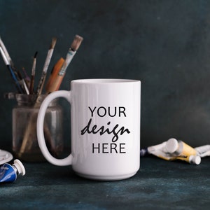 May include: White ceramic mug with a black inscription that reads "YOUR design HERE". The mug is sitting on a blue surface with paint tubes and brushes in the background.