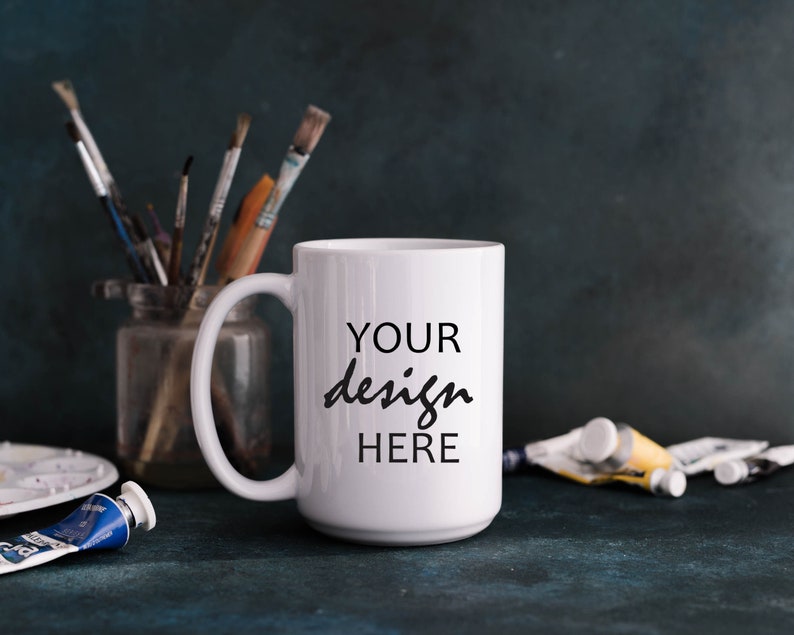 May include: White ceramic mug with a black inscription that reads "YOUR design HERE". The mug is sitting on a blue surface with paint tubes and brushes in the background.
