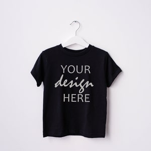 May include: A black t-shirt with short sleeves hanging on a white hanger. The shirt has the text "YOUR design HERE" printed on the front in a light gray color.
