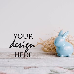 May include: A light blue ceramic bunny figurine sitting in a nest of straw on a whitewashed wooden surface. The text "YOUR design HERE" is written in black on the white background.