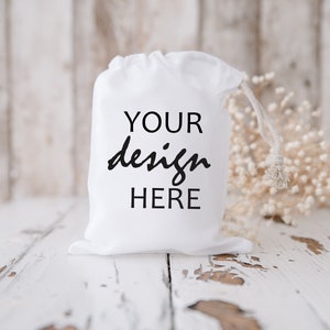 Könnte beinhalten: Ein weißer Beutel mit Kordelzug und dem Aufdruck "YOUR design HERE" auf der Vorderseite. Der Beutel liegt auf einer weißen Holzoberfläche.