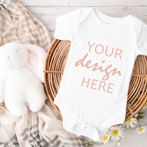 May include: White baby onesie with the text "YOUR design HERE" in pink on a wicker basket with a white stuffed bunny and white flowers.