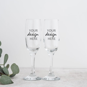 May include: Two clear glass champagne flutes with black text that reads "YOUR design HERE" on each flute.