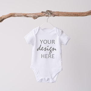 以下が含まれることがあります： 木製の枝に掛けられた白いベビーロンパース。前面に「YOUR design HERE」の文字が印刷されています。