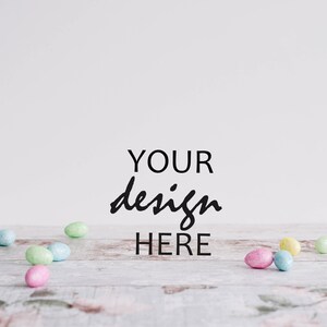 May include: A white background with the text "YOUR design HERE" in black cursive writing.  A light wood surface with pastel colored Easter eggs scattered across it.