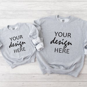 May include: Two grey sweatshirts with the text "YOUR design HERE" printed on the front. The sweatshirts are laid flat on a white wooden surface.