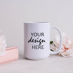May include: White ceramic mug with a black inscription that reads "YOUR design HERE" on a white background. The mug is sitting on a white surface with pink flowers and a pink book.