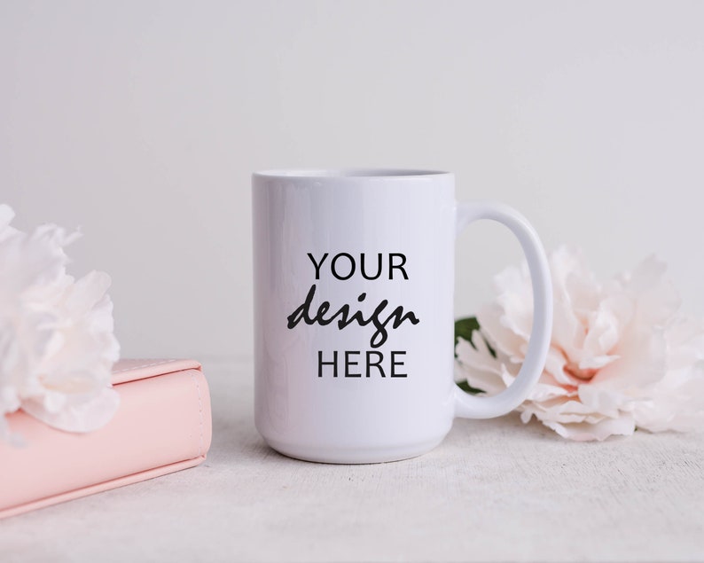 May include: White ceramic mug with a black inscription that reads "YOUR design HERE" on a white background. The mug is sitting on a white surface with pink flowers and a pink book.