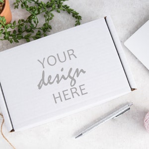 May include: White cardboard box with the text "YOUR design HERE" printed on the top. The box is sitting on a white surface with a pen, a ball of yarn, and a potted plant.
