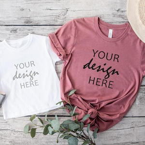 Puede incluir: Dos camisetas, una blanca y otra malva, colocadas sobre una superficie de madera. Ambas camisetas tienen el texto "YOUR design HERE" impreso en ellas.