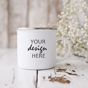 Könnte beinhalten: Weiße Emaille-Campingtasse mit schwarzer Aufschrift "YOUR design HERE" auf einer weiß getünchten Holzoberfläche mit weißen Blumen.