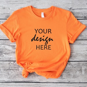 Op de afbeelding: Oranje T-shirt met opgerolde mouwen op een houten achtergrond. Het shirt heeft de tekst "YOUR design HERE" op de voorkant.