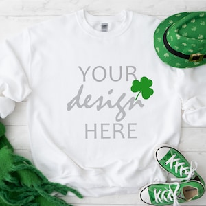 May include: White sweatshirt with the text "YOUR design HERE" in gray lettering and a green shamrock. The sweatshirt is laying on a white wooden surface with a green scarf, a green hat with shamrocks, and green and white sneakers.