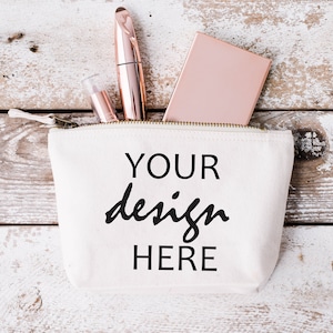 Può includere: Una pochette in tela bianca con cerniera con il testo "YOUR design HERE" stampato sul davanti. La pochette è aperta e contiene un compatto oro rosa, un mascara oro rosa e un lucidalabbra oro rosa.