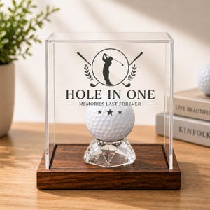 May include: A golf ball displayed in a clear acrylic case with a wooden base. The case features the text "HOLE IN ONE" and a silhouette of a golfer. The golf ball sits on a crystal stand, and the case is on a wooden surface.