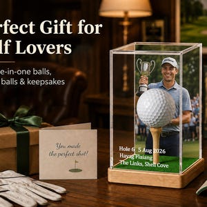 May include: A clear acrylic display case with a wooden base containing a golf ball on a tee and a photograph of a golfer holding a trophy. The case features white text: Hole 6, 5 Aug 2026, Hayag Flaiang, The Links, Shell Cove. A gift box and card are nearby.