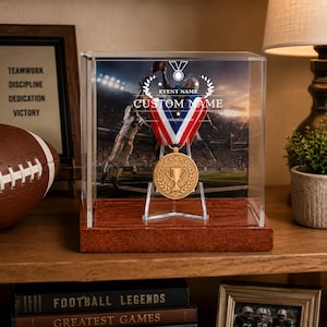 Könnte beinhalten: Eine goldfarbene Trophäenmedaille mit Lorbeerkranz-Design und Pokalsymbol, aufgehängt an einem rot-weiß-blauen Band in einer durchsichtigen Acryl-Vitrine auf einem Holzsockel, mit einem Hintergrundbild eines Football-Spiels.