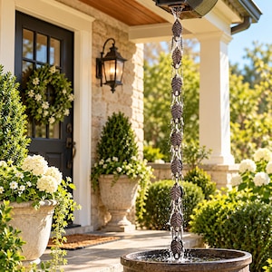 May include: A dark bronze rain chain featuring a series of interconnected metal leaf-shaped links that guide water from a roof gutter down into a circular basin, creating a vertical water feature in front of a residential entryway.