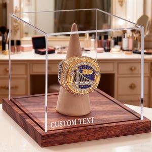 May include: A gold championship ring with a blue and gold design is displayed in a clear acrylic case. The ring rests on a tan cone, set on a dark wood base. The base features the text "CUSTOM TEXT".