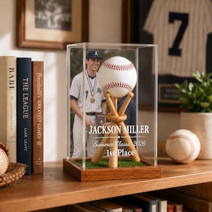 Op de afbeelding: Een doorzichtige acryl vitrinekast met een houten voet bevat een witte honkbal op een standaard van honkbalknuppels. Op de voorzijde staat de tekst JACKSON MILLER, Summer Classic 2026, 1st Place en een foto van een honkbalspeler afgebeeld.