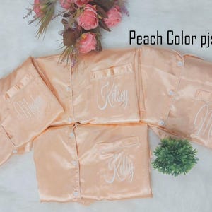 Può includere: Camicie da pigiama in raso color pesca con maniche lunghe, abbottonatura frontale e tasche sul petto. Ogni camicia presenta un ricamo in corsivo bianco con i nomi Meghan, Kelsey, Kelly e Mom. Il testo Peach Color pjs è visibile.