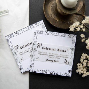 May include: Two memo pads labelled "Celestial Notes" are shown. Each pad has a white background with a black and white celestial border. The pads are 7.5cm x 10.5cm and contain 50 sheets of recycled paper. A candle holder and dried flowers are also in the image.