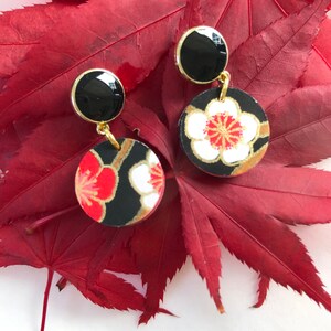 May include: A pair of dangle earrings with black and gold round charms. The charms feature a red and white floral pattern. The earrings are on a red background.