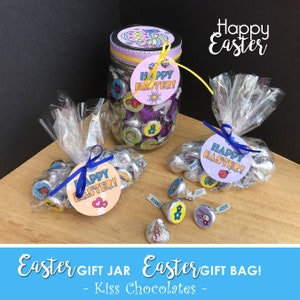 Peut inclure: Un bocal en verre transparent rempli de chocolats emballés dans du papier d'aluminium et décoré d'un couvercle violet et d'une étiquette "Joyeuses Pâques !". Deux sacs en plastique transparents remplis de chocolats emballés dans du papier d'aluminium et décorés d'un ruban bleu et d'étiquettes "Joyeuses Pâques !". Les sacs et le bocal sont posés sur une surface en bois.