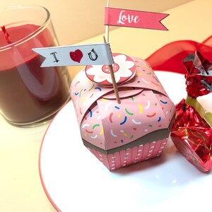 May include: A pink cupcake box with colorful sprinkles and a white flower on top. The box has a "Love" flag and an "I love you" flag attached to it.