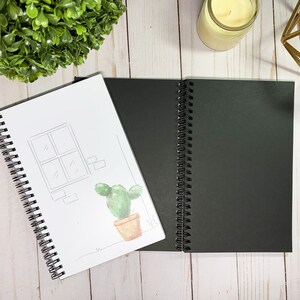 May include: Two spiral-bound notebooks, one with a black cover and the other with a white cover. The white cover has a watercolour illustration of a cactus in a pot, a window, and brick details. The notebooks are open.