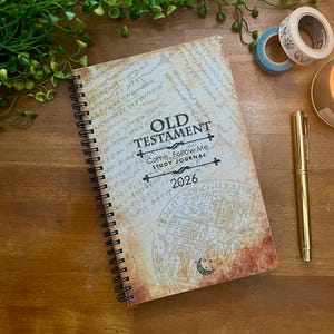 Puede incluir: Un diario de estudio con espiral con el texto "OLD TESTAMENT" y "Come, Follow Me Study Journal 2026" en la portada. La portada tiene un diseño vintage, similar al pergamino, con escritura e ilustraciones descoloridas. También se ven un bolígrafo dorado y rollos de cinta decorativa.